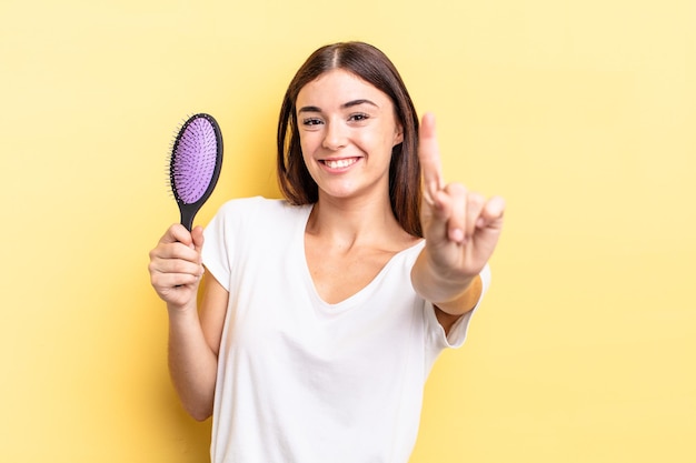 Young hispanic woman smiling proudly and confidently making number one. hair brush concept