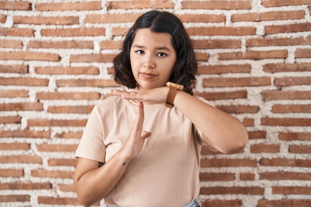 Young hispanic woman standing over bricks wall doing time out gesture with hands, frustrated and serious face