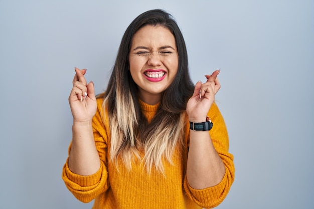 Young hispanic woman standing over isolated background gesturing finger crossed smiling with hope and eyes closed. luck and superstitious concept.