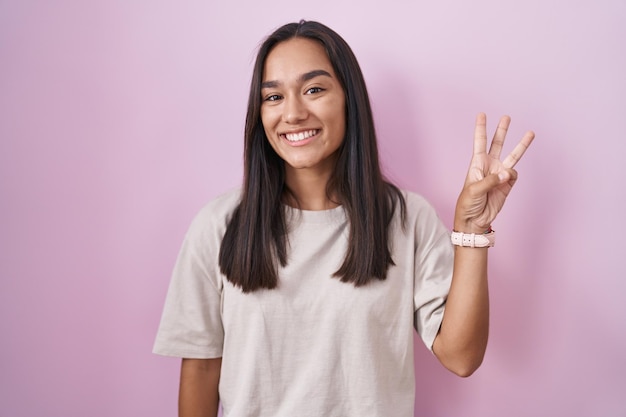 Young hispanic woman standing over pink background showing and pointing up with fingers number three while smiling confident and happy.