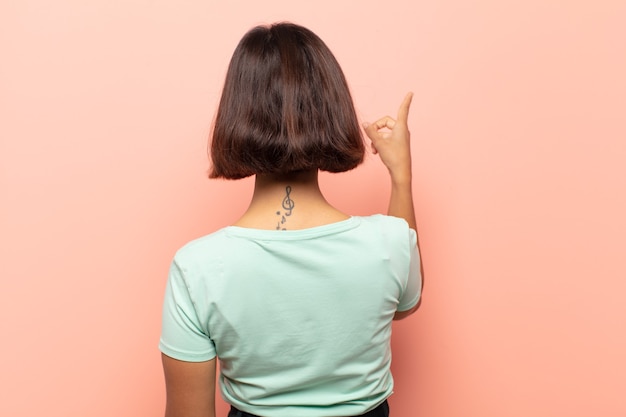 Photo young hispanic woman standing and pointing to object on copy space, rear view