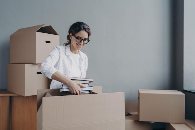 Young hispanic woman unpacking carton boxes and takes books out Female student moves to campus room Happy girl in glasses unloading luggage Relocation and entering to university