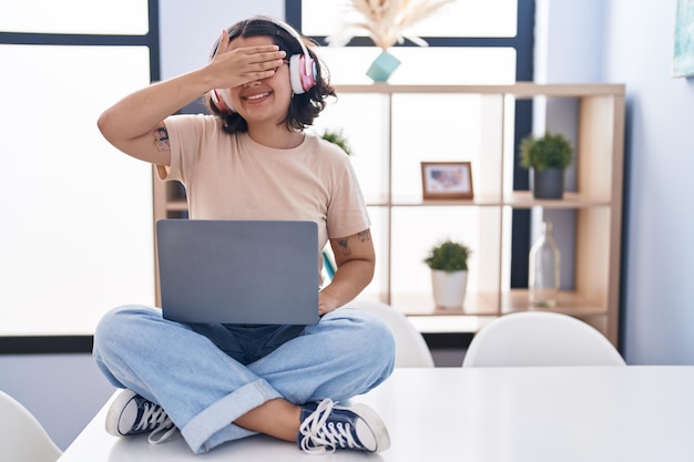 Young hispanic woman using laptop sitting on the table wearing headphones smiling and laughing with hand on face covering eyes for surprise blind concept