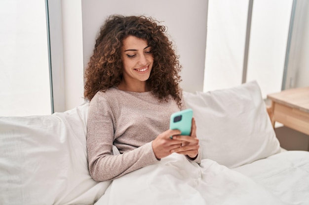 Young hispanic woman using smartphone sitting on bed at bedroom