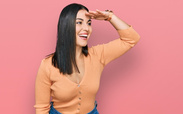 Young hispanic woman wearing casual clothes very happy and smiling looking far away with hand over head. searching concept.