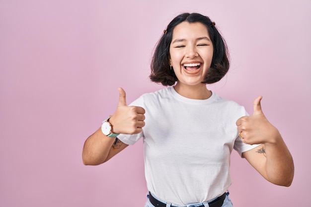 Young hispanic woman wearing casual white t shirt over pink background success sign doing positive gesture with hand, thumbs up smiling and happy. cheerful expression and winner gesture.