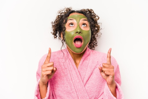 Young hispanic woman wearing a facial mask isolated on white background pointing upside with opened mouth