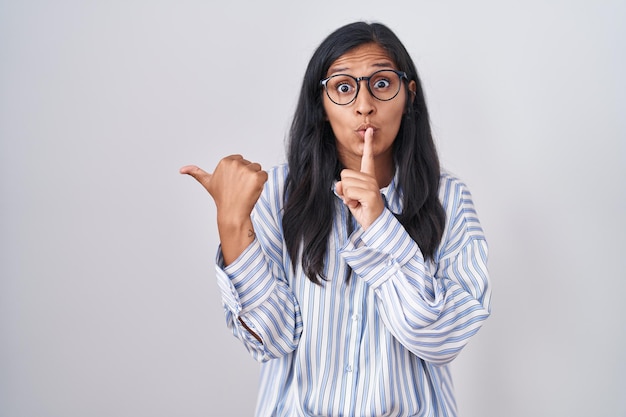 Young hispanic woman wearing glasses asking to be quiet with finger on lips pointing with hand to the side. silence and secret concept.