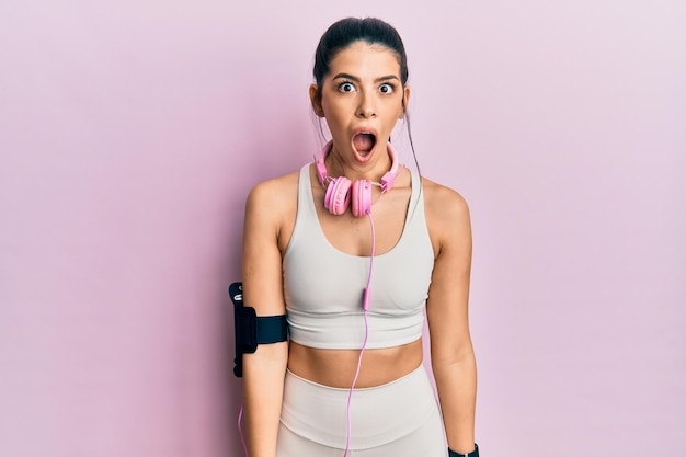 Young hispanic woman wearing gym clothes and using headphones afraid and shocked with surprise and amazed expression fear and excited face