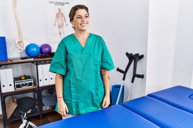 Young hispanic woman wearing physiotherapist uniform standing at clinic looking away to side with smile on face, natural expression. laughing confident.