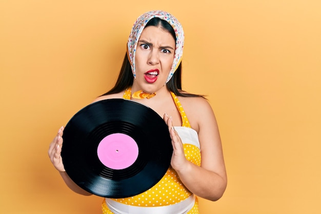 Young hispanic woman wearing pin up style holding vinyl disc in shock face looking skeptical and sarcastic surprised with open mouth