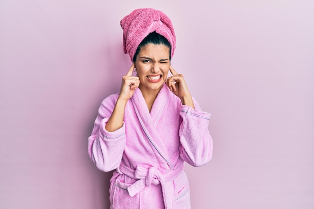 Young hispanic woman wearing shower towel cap and bathrobe covering ears with fingers with annoyed expression for the noise of loud music deaf concept