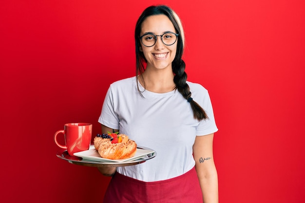 Young hispanic woman wearing waitress apron holding tray with breakfast looking positive and happy standing and smiling with a confident smile showing teeth