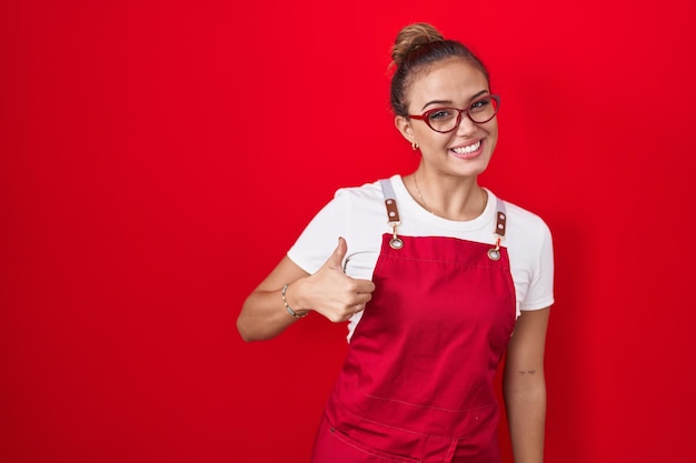 Young hispanic woman wearing waitress apron over red background doing happy thumbs up gesture with hand. approving expression looking at the camera showing success.