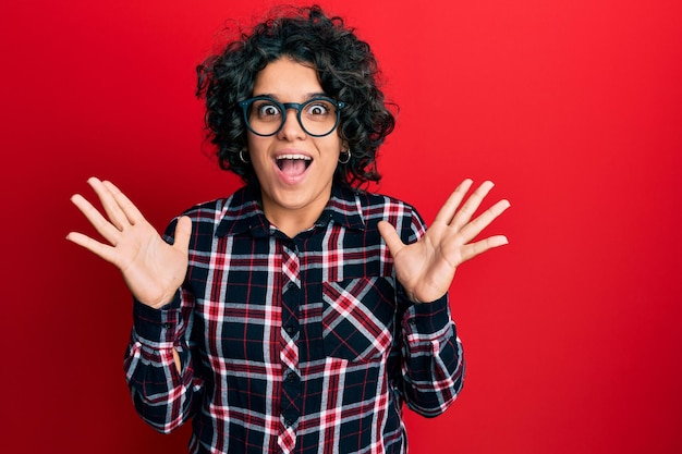 Young hispanic woman with curly hair wearing casual clothes and glasses celebrating crazy and amazed for success with arms raised and open eyes screaming excited winner concept