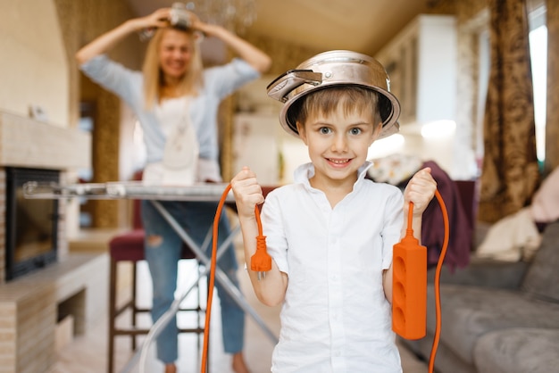 Young housewife irons the clothes, little boy dabbles with electricity. Woman doing housework at home. Female person with her playful son in the house