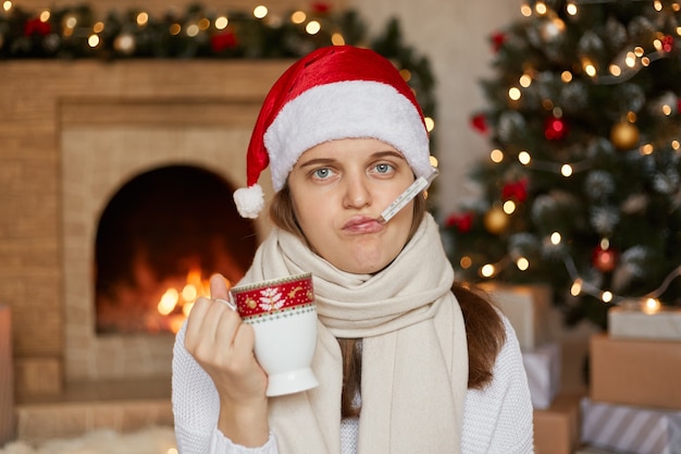 Young ill woman in santa hat, wrapped in scarf, measuring temperature and holding cup of warming drink,, being sick on Christmas holiday, posing in room with new Year decorations.