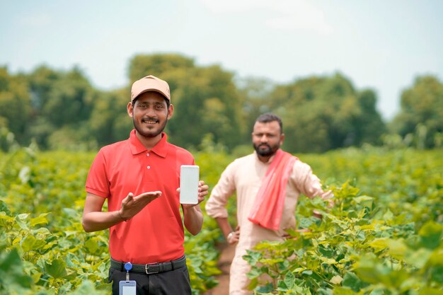 Young indian agronomist or banker showing smartphone with farmers at agriculture field.