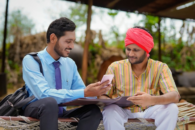 Young indian banker or agronomist showing some detail to farmer in smartphone.