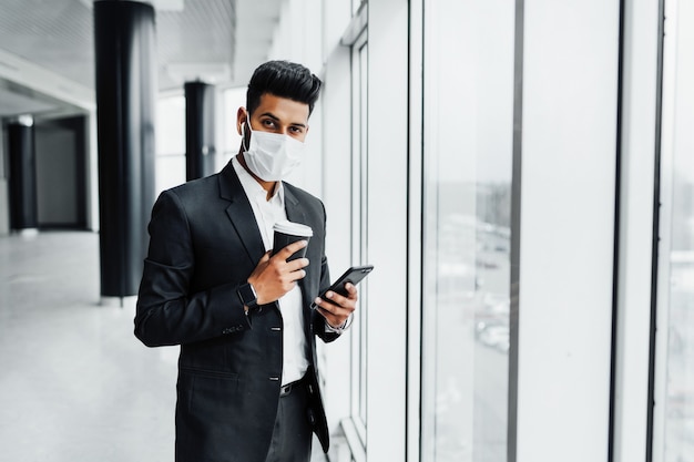 A young Indian businessman in a protective mask by the window, he holds his phone in his hands and looks at the camera.