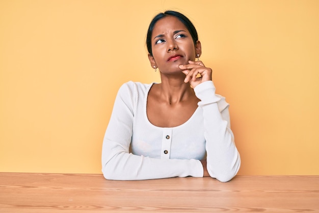 Young indian girl wearing casual clothes sitting on the table serious face thinking about question with hand on chin thoughtful about confusing idea