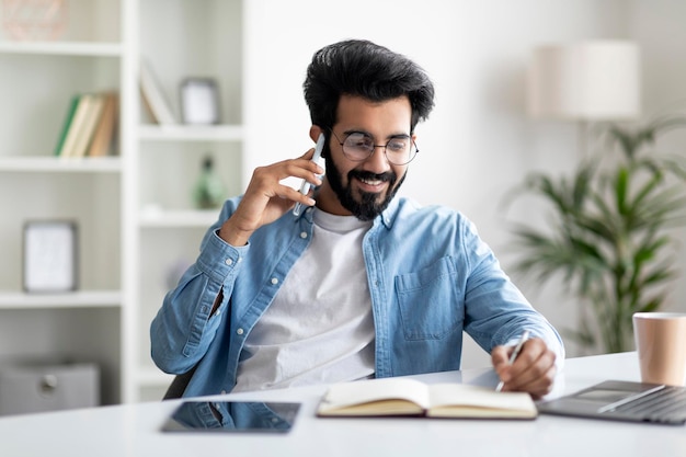 Young indian male entrepreneur talking on cellphone and taking notes in office