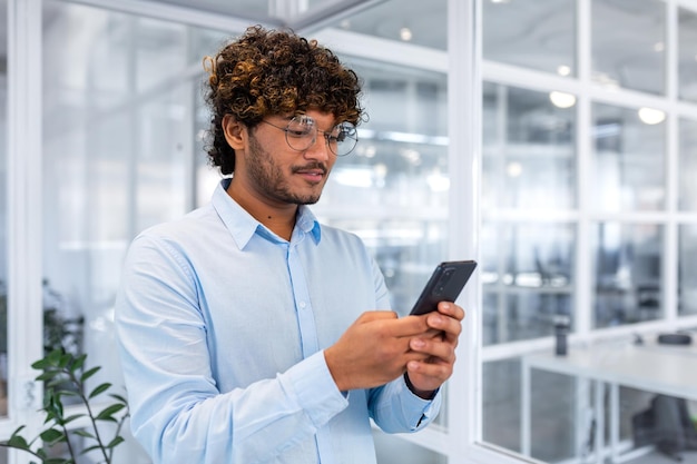 A young indian man is standing in the office and using the phone dialing messages texting online