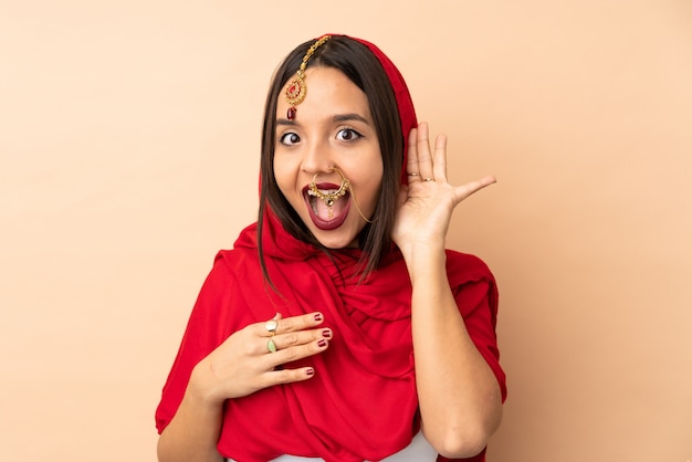 Young Indian woman on beige wall listening to something by putting hand on the ear