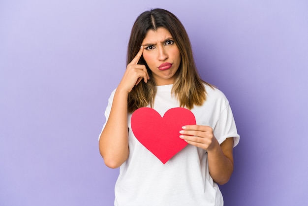 Young indian woman holding a valentines day heart isolated showing a disappointment gesture with forefinger.