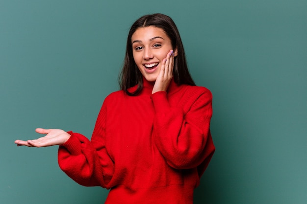 Young Indian woman isolated on blue background holds copy space on a palm, keep hand over cheek. Amazed and delighted.