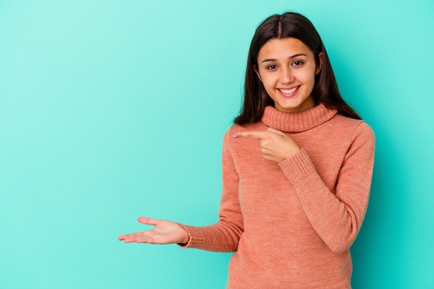 Young Indian woman isolated on blue wall excited holding a copy space on palm.