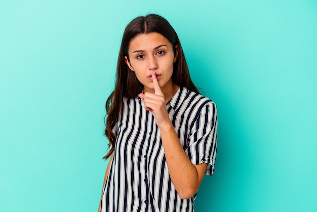 Young Indian woman isolated on blue wall keeping a secret or asking for silence.