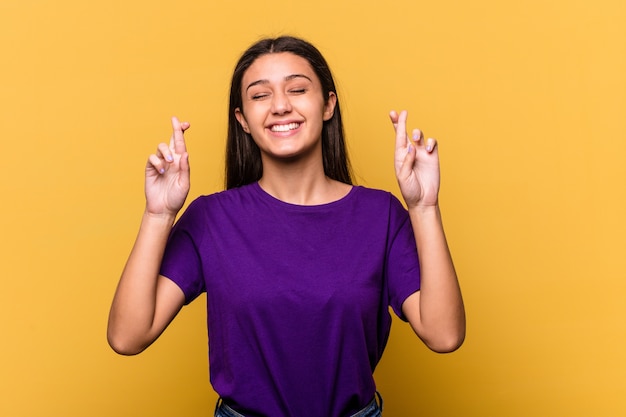 Young Indian woman isolated on yellow wall crossing fingers for having luck