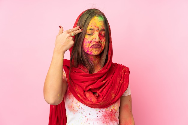 Young Indian woman with colorful holi powders on her face on pink wall with problems making suicide gesture