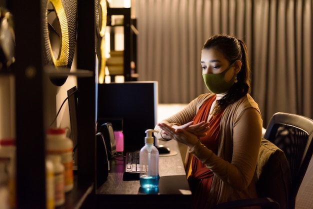 Young Indian woman with mask using hand sanitizer and rubbing hands together while working from home at night