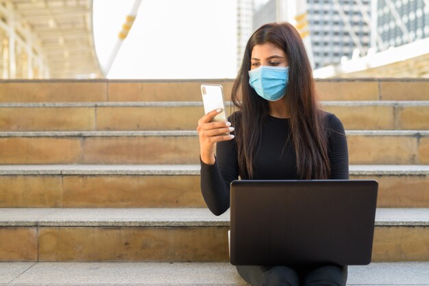 Young Indian woman with mask using laptop and phone by the stairs in city