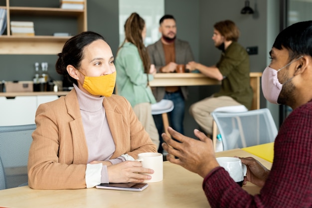 Young intercultural designers or managers in casualwear and protective masks having coffee and discussing working plans at break