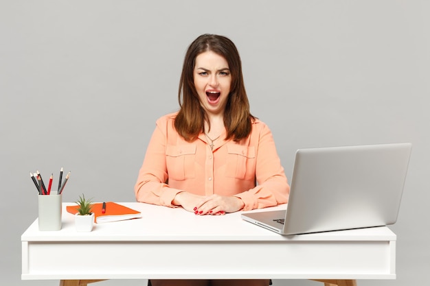 Young irritated woman in pastel casual clothes scareaming swearing sit work at white desk with pc laptop isolated on gray background. Achievement business career lifestyle concept. Mock up copy space.