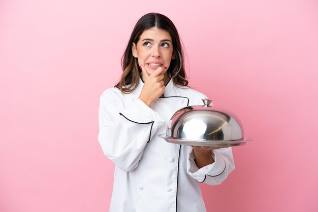 Young Italian chef woman holding tray with lid isolated on pink background having doubts and thinking