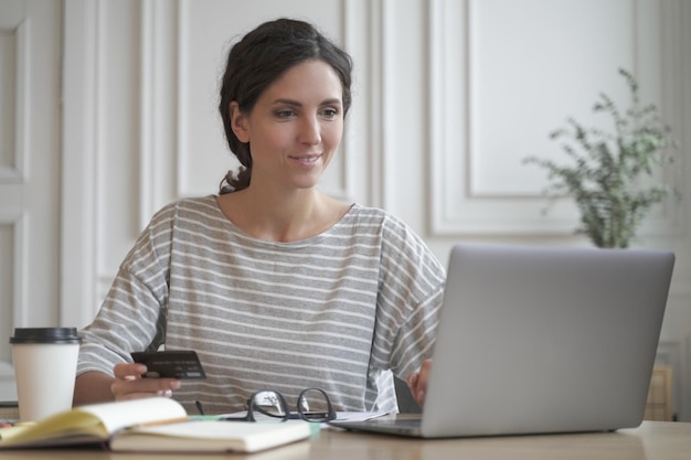 Young Italian female freelancer holding credit card using laptop while making payments online