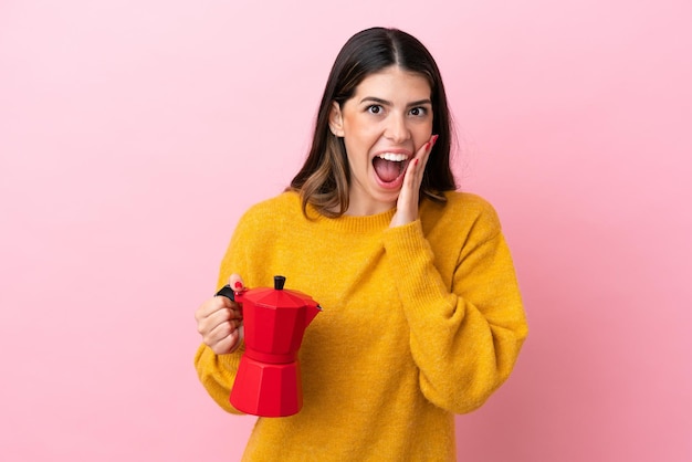 Young Italian woman holding a coffee maker isolated on pink background with surprise and shocked facial expression