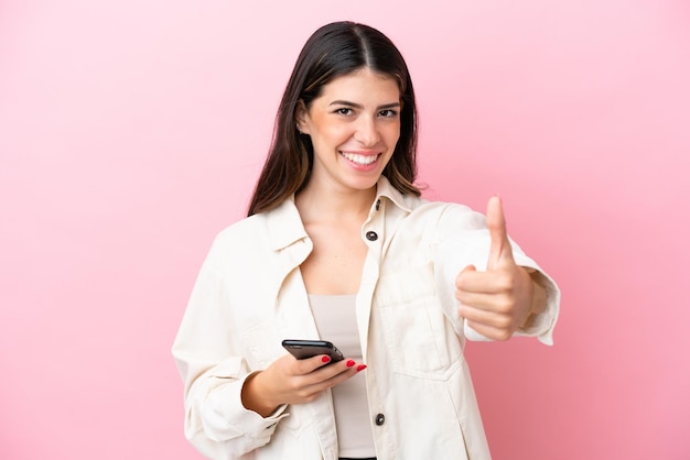 Young Italian woman isolated on pink background using mobile phone while doing thumbs up