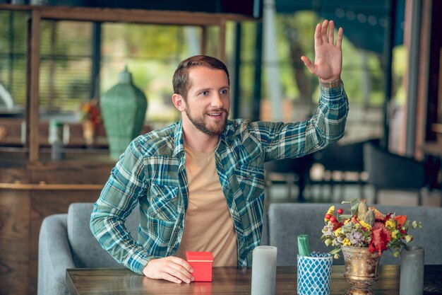 Young joyful bearded man with gift raising his hand sitting at table in summer cafe