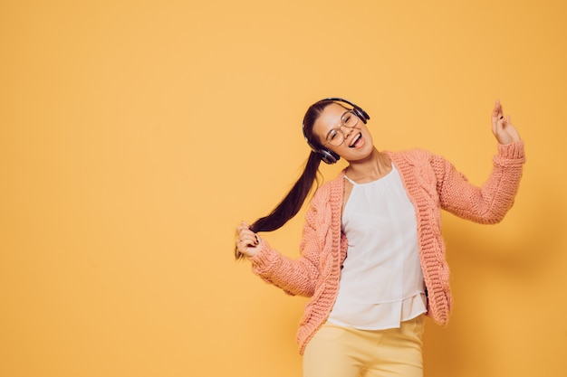 Young joyful brunette in glasses and headphones on her head  dressed in pink sweater white blouse and yellow pants enjoying the music, singing and dancing over yellow background with copy space.