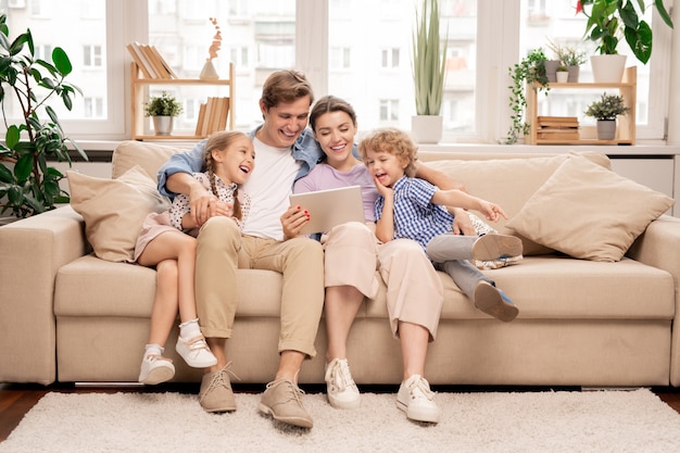 Young joyful casual family of two kids and couple sitting on sofa and watching funny video