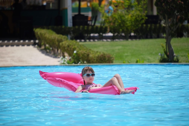 Young joyful child girl having fun swimming on inflatable air mattress in swimming pool with blue water on warm summer day on tropical vacations Summertime activities concept