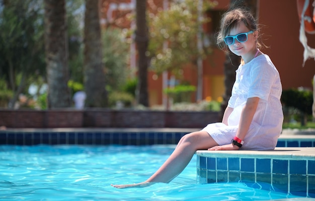 Young joyful child girl resting on swimming pool side with clear blue water on sunny summer day Tropical vacations concept