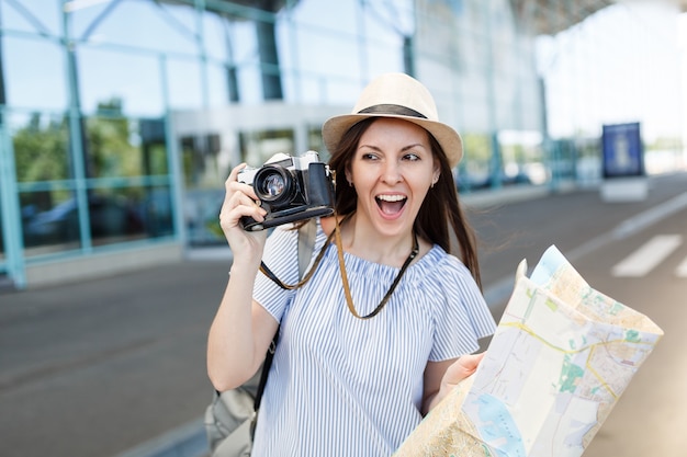 Young joyful traveler tourist woman in hat holding retro vintage photo camera, paper map at international airport