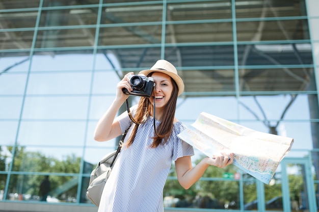 Young joyful traveler tourist woman take pictures on retro vintage photo camera holding paper map at international airport