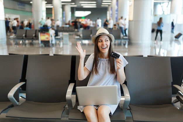 Young joyful traveler tourist woman working on laptop hold credit card spreading hands, wait in lobby hall at international airport